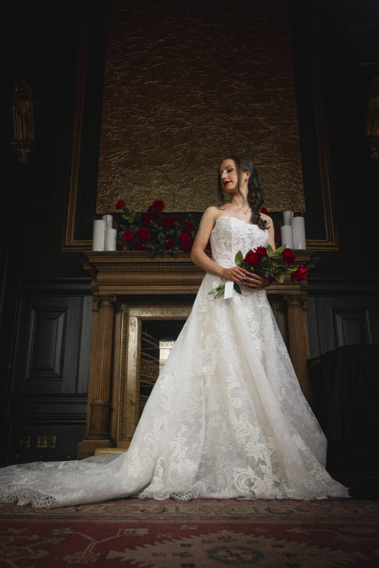 Bride in a lace wedding gown holding red roses, standing before the ornate fireplace at Hutchesons' Hall, Glasgow's Merchant City wedding venue